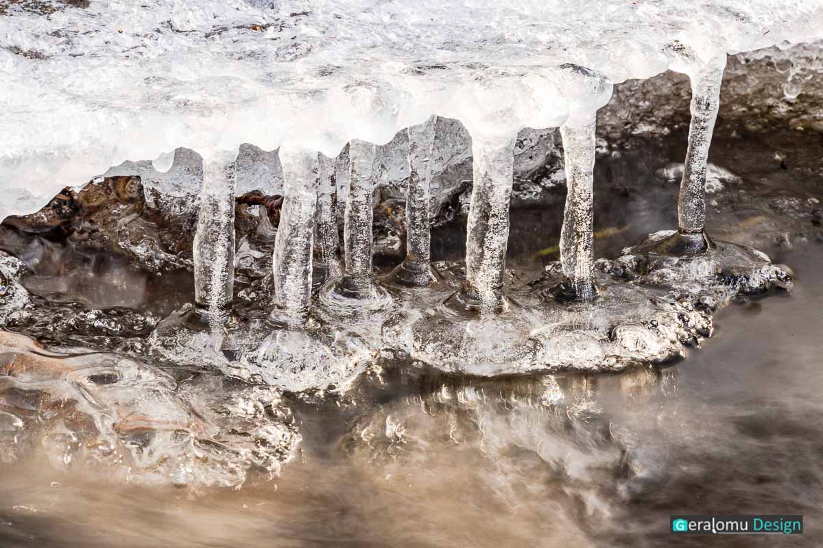 Kreative Fotografie: Dicke Eiszapfen am Bachufer mit interessanter Wasserströmung in Makroaufnahme.