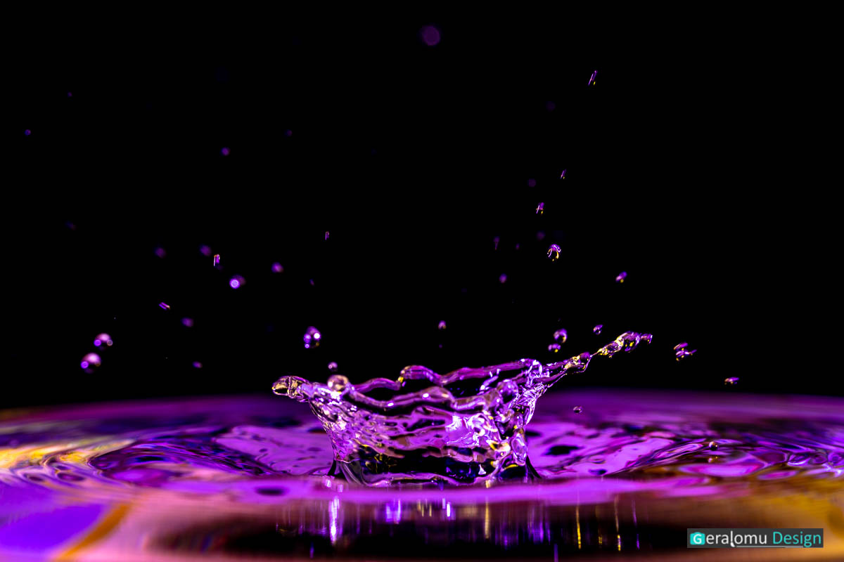 Makro: Sprühende blau und lila gefärbte Wassertröpfchen nach Aufprall eines Tropfens auf eine Wasseroberfläche