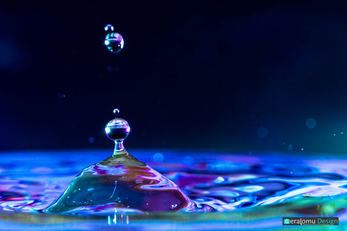 Wassertropfenfotografie Makro: Zwei dicke blau- und lilafarbige schweben nach dem Aufprall eines Wassertropfens über der Wasseroberfläche