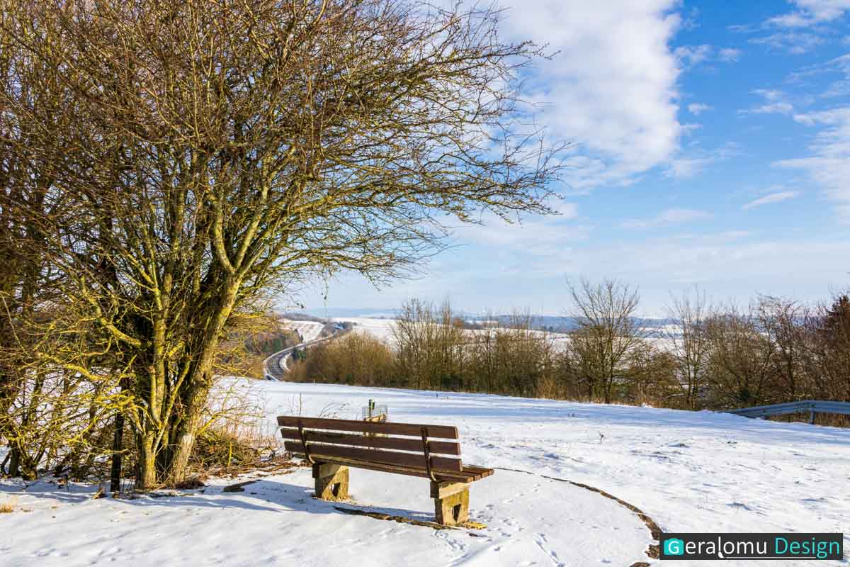 Diese Landschaftsfotografie zeigt eine leere Sitzbankin schneebedeckter Winterlandschaft in der Umgebung des Ortes Ellwerath im Eifelkreis Bitburg-Prüm.